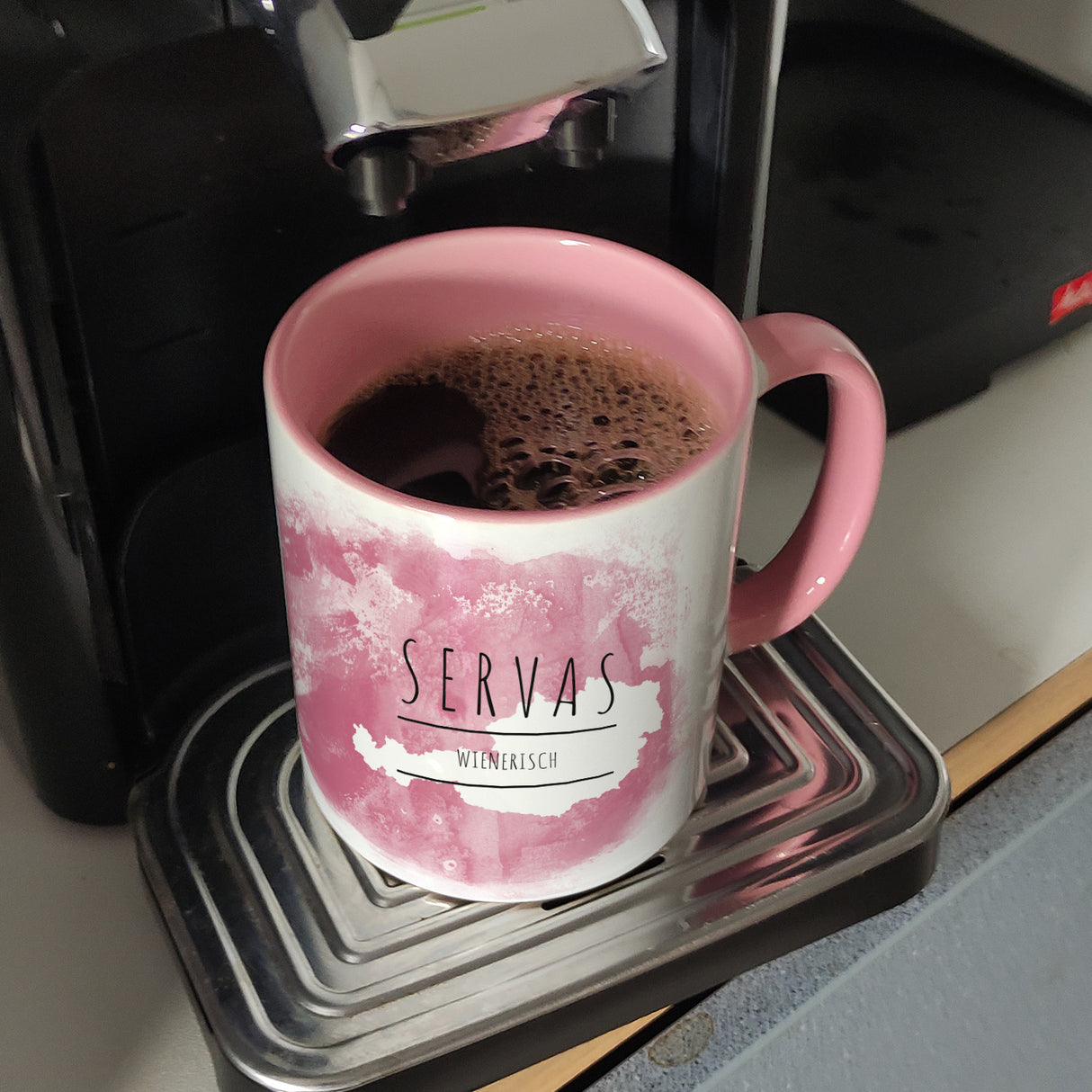 Hallo auf Wienerisch Servas lustiger Kaffeebecher mit rosaem Hintergrund
